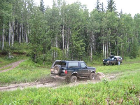 1995 Ford Bronco - Two SUVs navigate a muddy trail surrounded by a forest.