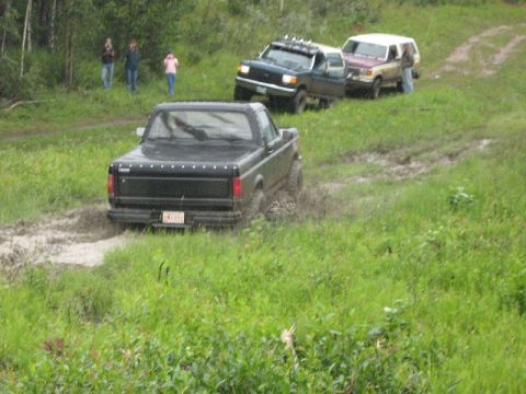 1995 Ford Bronco - Multiple trucks navigate a muddy off-road course with onlookers present.