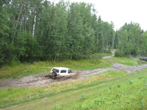 1995 Ford Bronco - A white SUV navigates a muddy track, kicking up spray as it gets stuck.