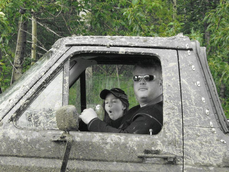 Mud-covered off-road vehicle interior with a man wearing sunglasses and a child in a baseball cap