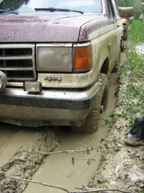 1995 Ford Bronco - A maroon and white Ford F-series truck is stuck in deep mud on a trail, with another vehicle visible behind it.