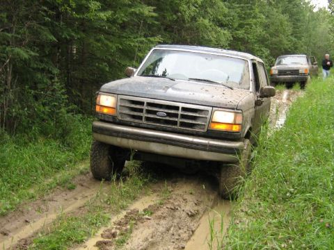 1995 Ford Bronco - Two Ford Broncos navigate a muddy trail through a wooded area.