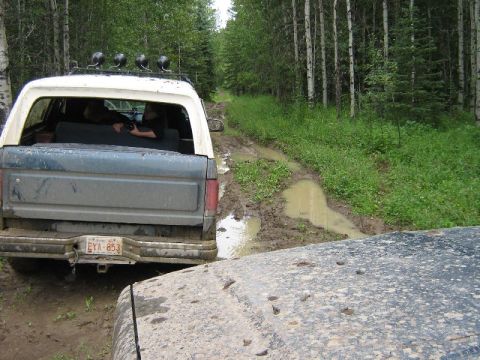 1995 Ford Bronco - A 1995 Ford Bronco navigates a muddy trail through a forest, with another vehicle visible in the foreground.