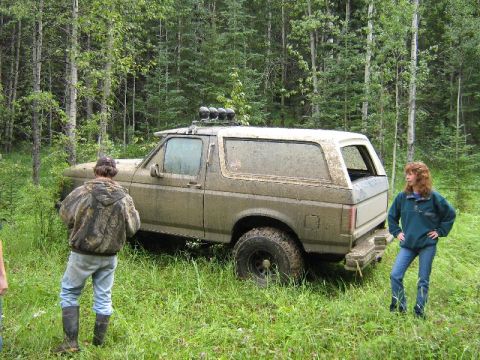 1995 Ford Bronco - A muddy Ford Bronco is parked off-road in a wooded area with two people observing.
