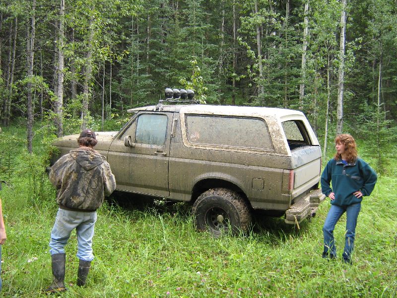 Mud-covered Ford Bronco with white hardtop and roof light bar parked in tall grass by woods