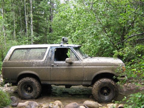1995 Ford Bronco - A muddy Ford Bronco navigates a water obstacle in a wooded area, its off-road lights visible.