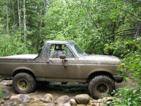 1995 Ford Bronco - A muddy Ford Bronco navigates a rocky stream bed surrounded by dense forest foliage.