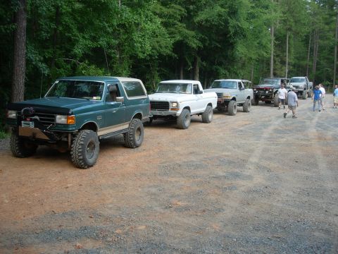 1995 Ford Bronco - A line of off-road vehicles, including a green 1995 Ford Bronco, are parked on a dirt path surrounded by trees.
