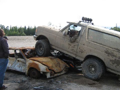 1995 Ford Bronco - A large, mud-splattered SUV is parked on top of a heavily damaged and burnt car.