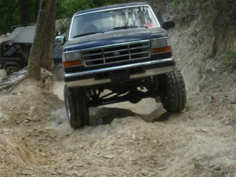 1995 Ford Bronco - A dark blue Ford Bronco navigates a challenging off-road trail with a lifted suspension and large tires.