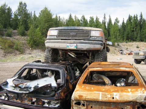 1995 Ford Bronco - A lifted 1995 Ford Bronco is perched atop two destroyed cars in a junkyard setting.
