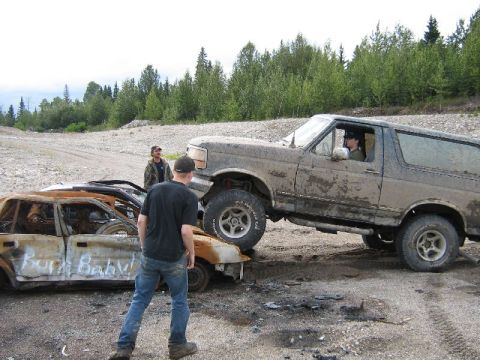 1995 Ford Bronco - A muddy SUV is positioned on top of a heavily damaged and burnt car, with two people observing the scene.