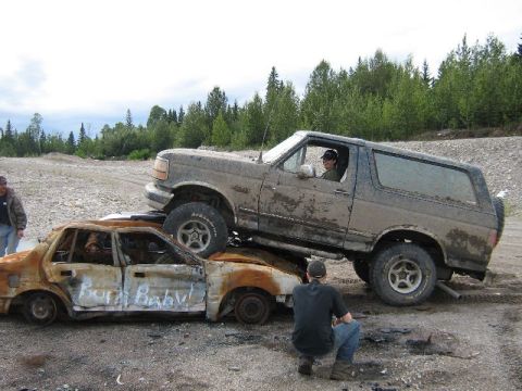 1995 Ford Bronco - A Ford Bronco is driven over a heavily damaged and burnt sedan in a dirt lot.