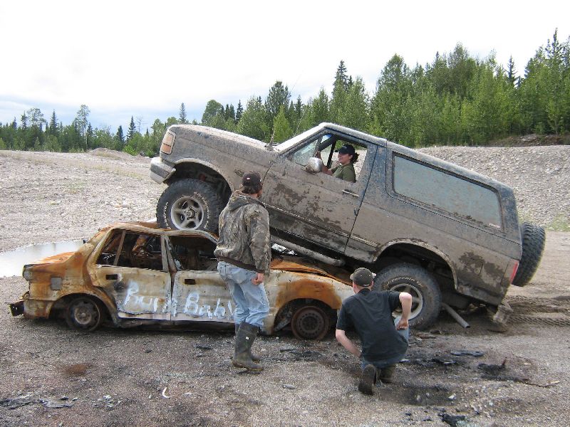 1995 Ford Bronco driving over a burnt car with "Burn Baby" written on its side