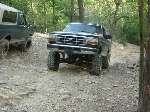 1995 Ford Bronco - Two Ford Broncos navigate a rocky off-road trail in a wooded area.