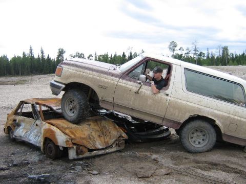 1995 Ford Bronco - A Ford Bronco is shown driving over a heavily damaged and burnt car in a dirt lot.