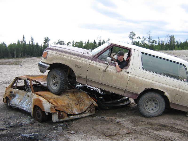 Ford Bronco driving over a burnt-out car. Driver is visible