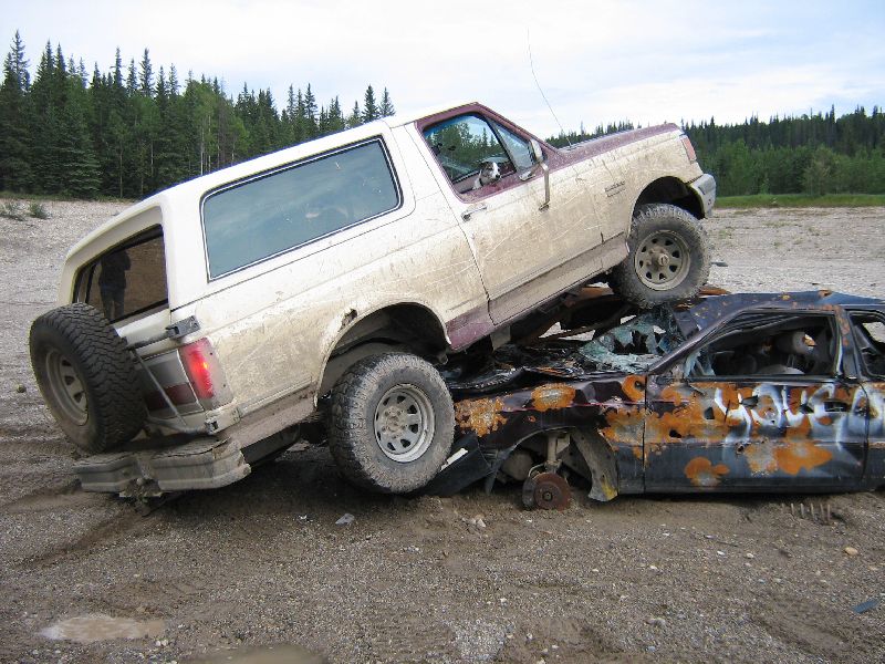 Mud-splattered white and maroon Ford Bronco on top of a rusted, wrecked car