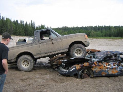1995 Ford Bronco - A muddy 1995 Ford Bronco is positioned atop two heavily damaged cars in a junkyard setting.
