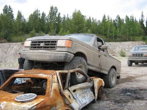 1995 Ford Bronco - A lifted Ford Bronco is positioned atop a destroyed vehicle, with another truck in the distance.