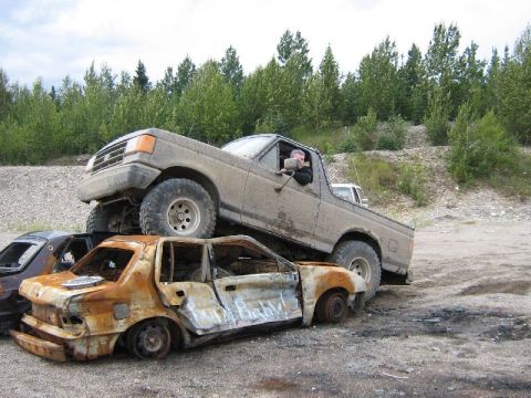 1995 Ford Bronco - A pickup truck is driven onto two heavily damaged cars in an outdoor junkyard setting.