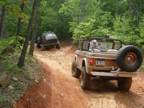 1995 Ford Bronco - Two off-road vehicles navigate a steep, rocky trail through a dense forest.