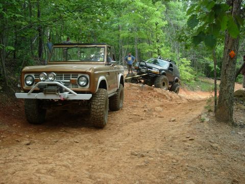 1995 Ford Bronco - A brown convertible SUV with a winch and off-road tires navigates a steep dirt trail, with another vehicle being towed behind.