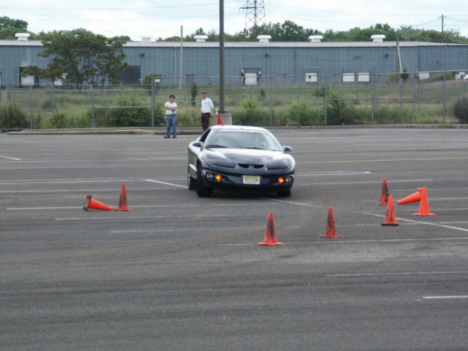 Dark blue Pontiac Firebird driving through cones in a parking lot. Two people stand in the background