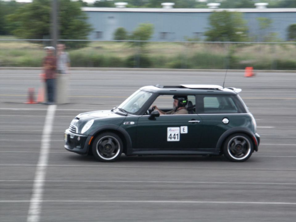 Dark green Mini Cooper with racing numbers 441 and E on the door, driving on a track. Driver wears a helmet