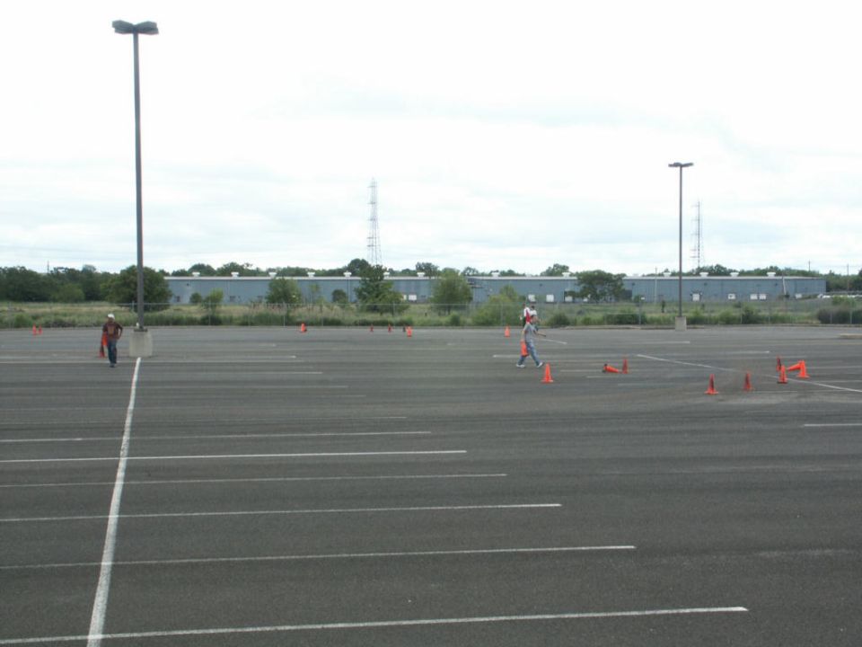 Empty asphalt parking lot with white lines, orange cones, two people, and a distant industrial building under a cloudy sky