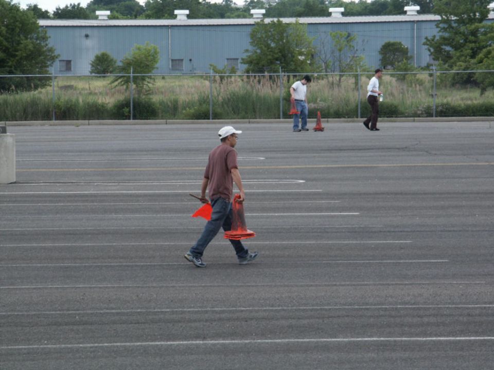 Man in maroon shirt and white hat carries orange traffic cone and baton across empty parking lot