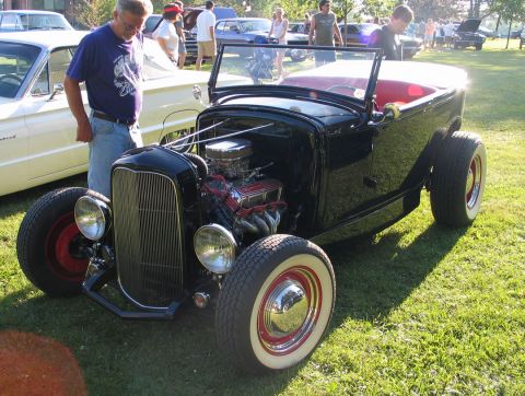 1994 Ford Bronco - A black hot rod with a prominent grille and exposed engine sits on a grassy lawn at a car show.