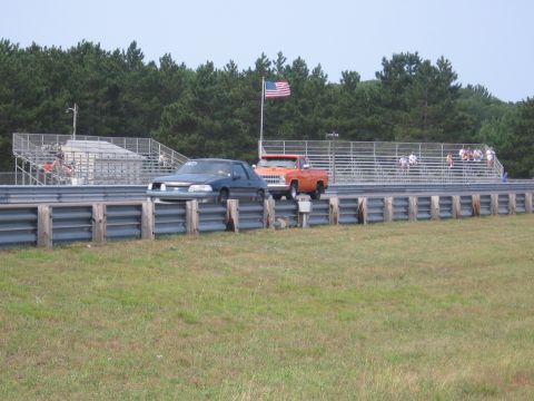 1989 Ford Mustang - Two vehicles race down a drag strip with an American flag flying overhead.