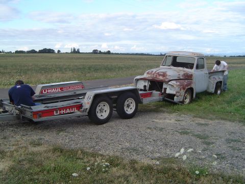 1955 Ford F-100 - A weathered pickup truck is being loaded onto a U-Haul trailer, suggesting a move or transport for restoration.