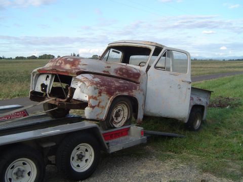 1955 Ford F-100 - A heavily rusted 1955 Ford F-100 truck is being transported on a trailer, missing its front grille and headlights.