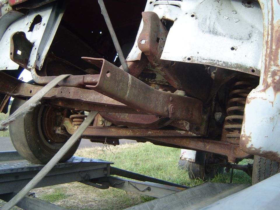 Underside of a rusty white classic pickup being winched onto a trailer, frame and coil spring visible