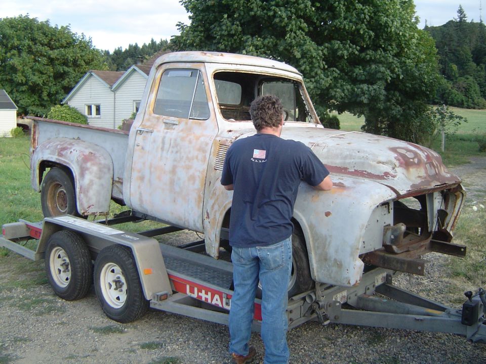 Rusty 1950s Ford stepside pickup shell strapped to a U-Haul trailer in a grassy yard