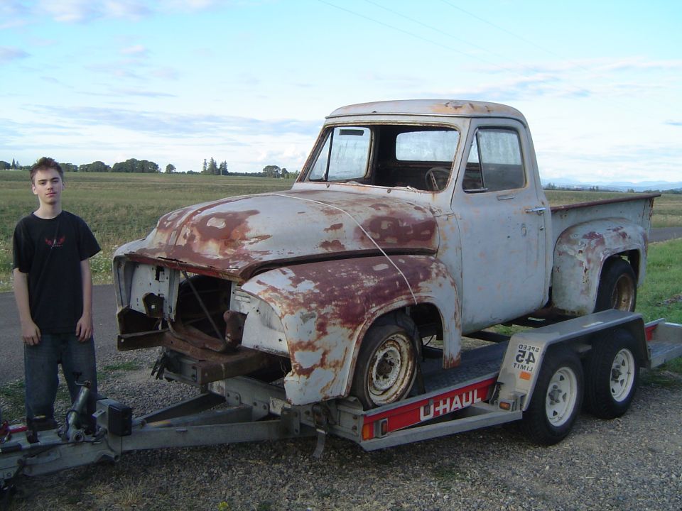 Guy stands next to a rusty 1955 Ford F-100 pickup truck on a U-Haul trailer