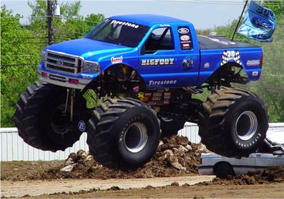 1996 Ford Bronco - A blue Bigfoot monster truck is airborne over a crushed car, with a Ford flag in the background.