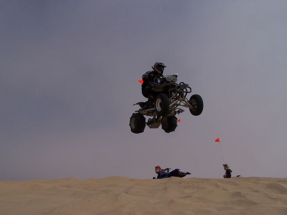 Quad bike rider airborne over sand dunes with two people on the ground