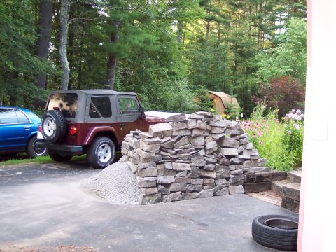 2001 Ford Crown Victoria - A red Jeep Wrangler is parked next to a partially constructed stone wall, with a blue car visible to its left.
