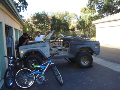 1971 Ford Bronco - Two men work on a disassembled 1971 Ford Bronco in a driveway, with bicycles nearby.