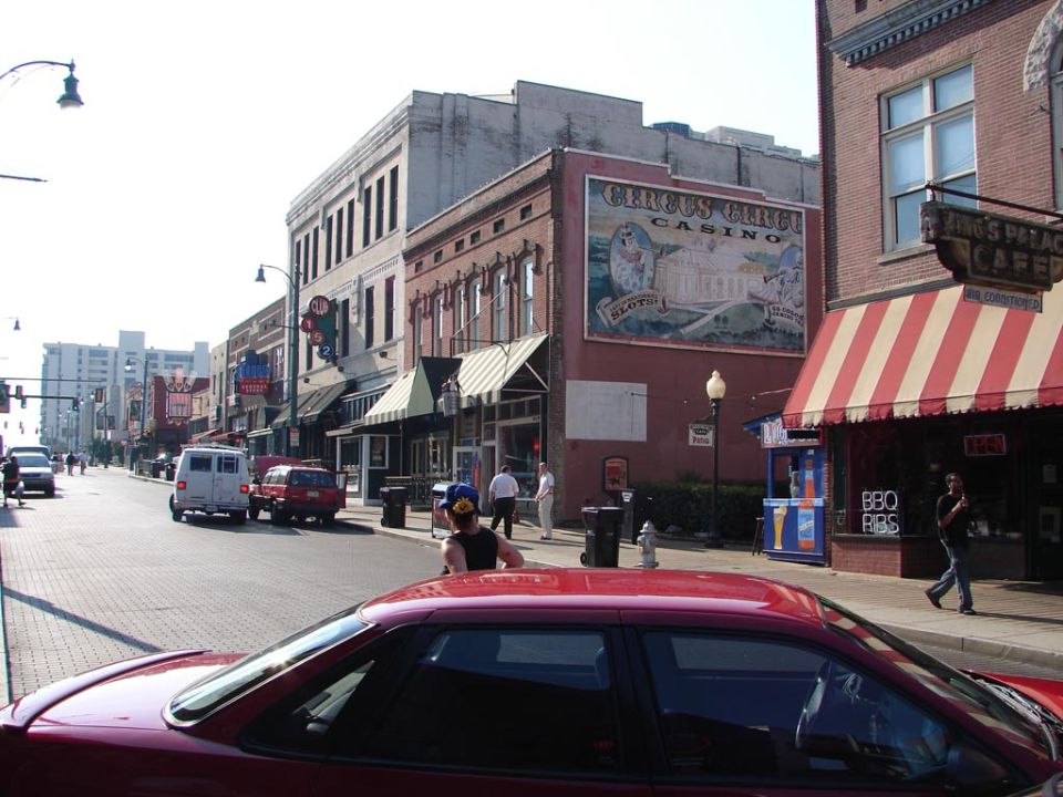 Street view with buildings, a red car in foreground, and a van and SUV on the road