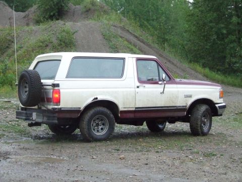 1990 Ford Bronco - A two-tone white and maroon 1990 Ford Bronco is parked on a dirt road with a spare tire mounted on the rear.