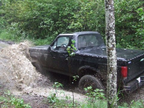 1990 Ford Bronco - A black pickup truck navigates a muddy path, kicking up water and dirt amidst a forest setting.