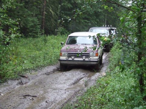 1990 Ford Bronco - A convoy of SUVs navigates a challenging muddy track through a dense forest.