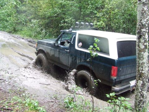 1990 Ford Bronco - An off-road vehicle navigates a challenging muddy path surrounded by dense foliage.