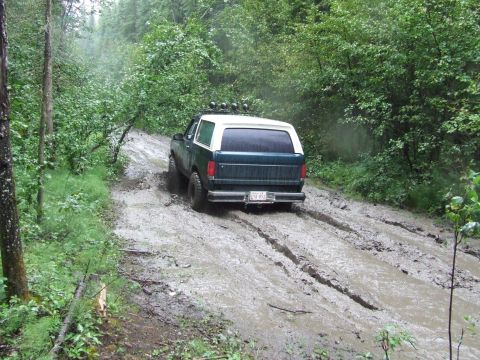 1990 Ford Bronco - A 1990 Ford Bronco navigates a challenging muddy path through a dense forest.