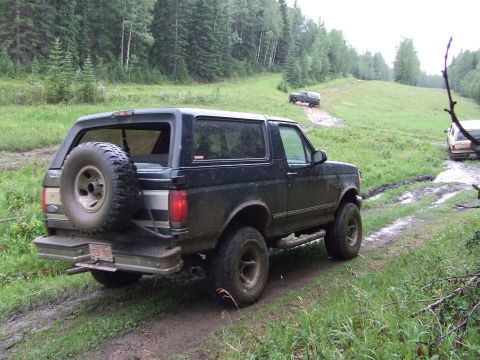 1990 Ford Bronco - A dark green Ford Bronco navigates a muddy off-road trail, with another vehicle visible further up the path.