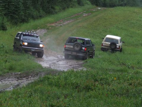 1990 Ford Bronco - Three SUVs navigate a muddy track on a grassy incline, with the lead vehicle being a dark green Ford Bronco.